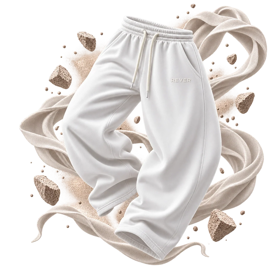 White sweatpants with a drawstring waist and a subtle 'REVER' logo on the left thigh, presented with stylized beige fabric swirls and small rocks on a white background.