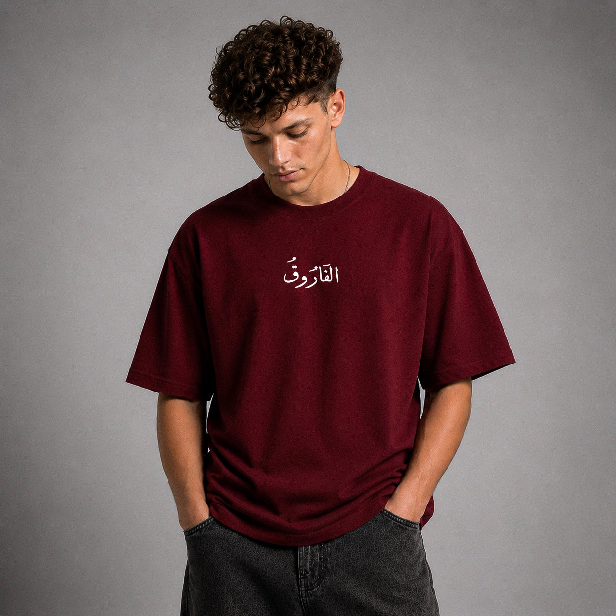 A man wearing a burgundy t-shirt with white Arabic calligraphy on the chest, paired with dark gray jeans, standing against a neutral gray studio background.