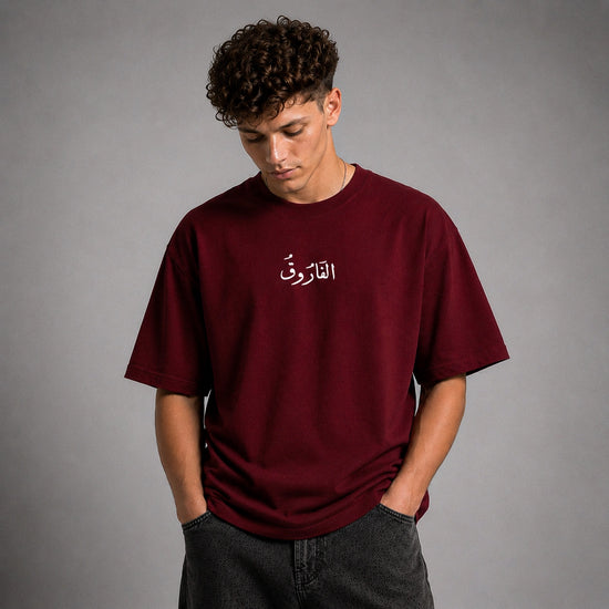 A man wearing a burgundy t-shirt with white Arabic calligraphy on the chest, paired with dark gray jeans, standing against a neutral gray studio background.