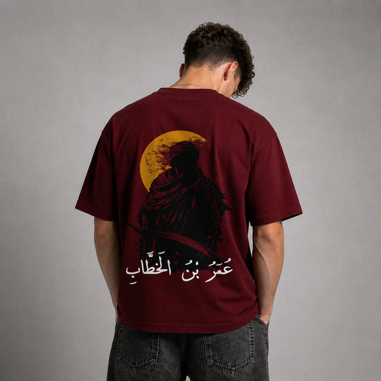 A man viewed from behind wearing a maroon t-shirt with a large graphic print of a hooded figure in front of a yellow full moon, with white Arabic text below. He is wearing dark gray jeans against a neutral gray studio background.