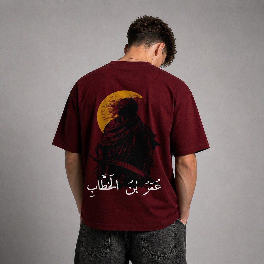 A man viewed from behind wearing a maroon t-shirt with a large graphic print of a hooded figure in front of a yellow full moon, with white Arabic text below. He is wearing dark gray jeans against a neutral gray studio background.