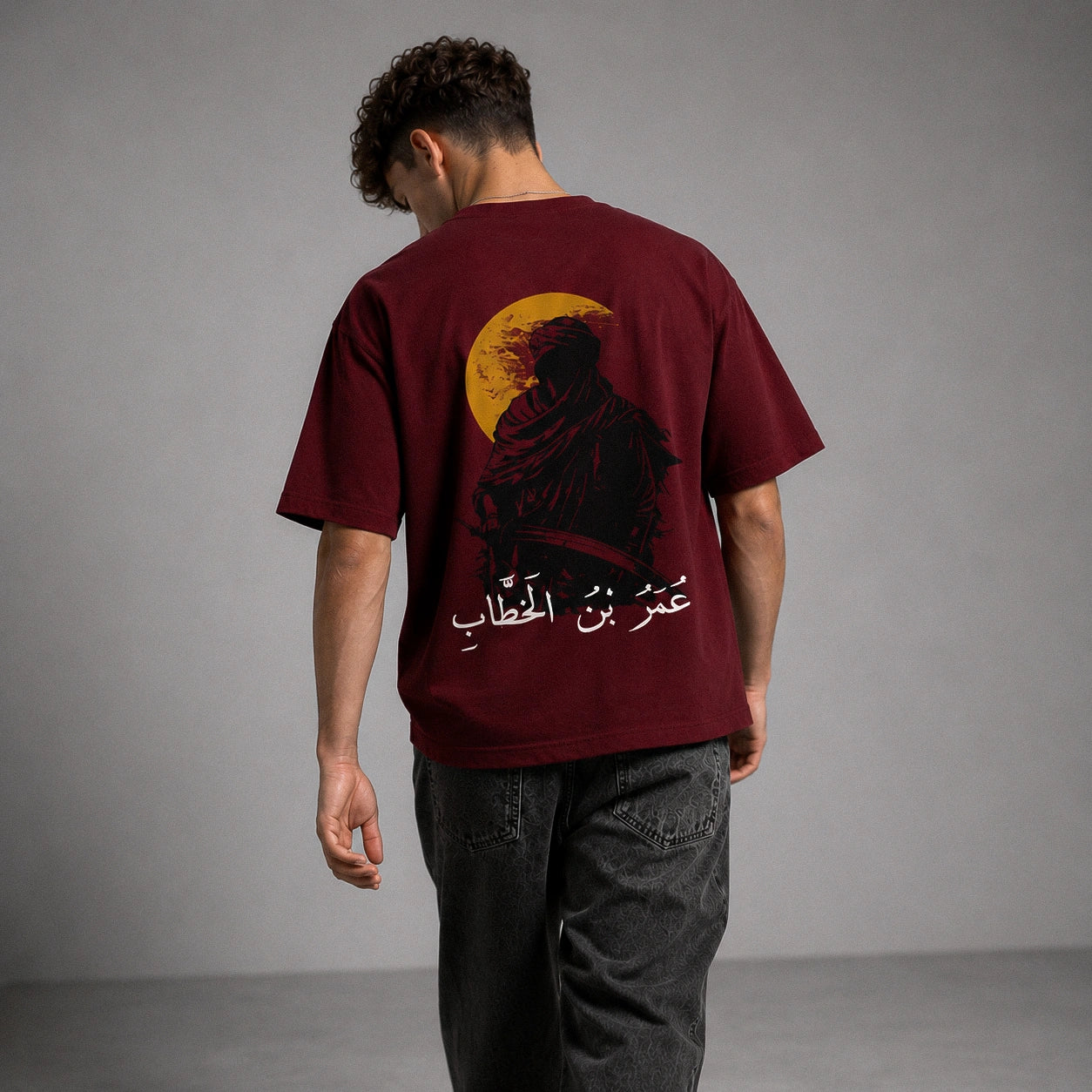 A man viewed from behind wearing a maroon t-shirt with a large graphic print of a robed figure in front of a yellow full moon and white Arabic text, paired with dark gray jeans against a gray studio background.