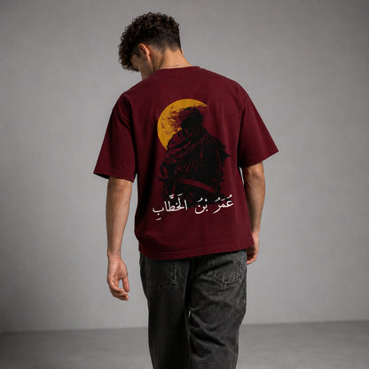 A man viewed from behind wearing a maroon t-shirt with a large graphic print of a robed figure in front of a yellow full moon and white Arabic text, paired with dark gray jeans against a gray studio background.