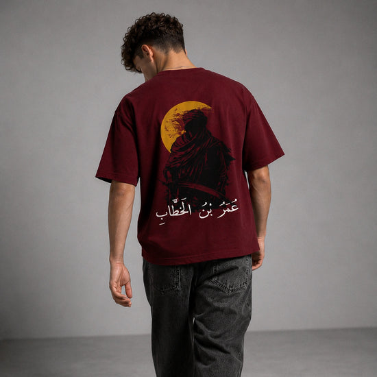 A man viewed from behind wearing a maroon t-shirt with a large graphic print of a robed figure in front of a yellow full moon and white Arabic text, paired with dark gray jeans against a gray studio background.