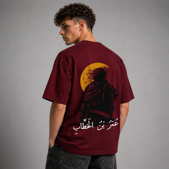 A man wearing a maroon t-shirt from the back, featuring a graphic print of a silhouette against a yellow moon with white Arabic text below it, set against a plain gray studio background.