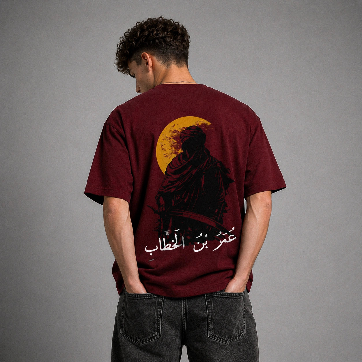 A man viewed from behind wearing a maroon t-shirt with a large graphic print on the back featuring a black silhouette, a yellow crescent moon, and white Arabic text, paired with dark gray jeans against a gray studio background.