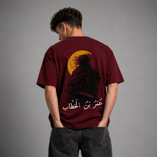 A man viewed from behind wearing a maroon t-shirt with a large graphic print on the back featuring a black silhouette, a yellow crescent moon, and white Arabic text, paired with dark gray jeans against a gray studio background.