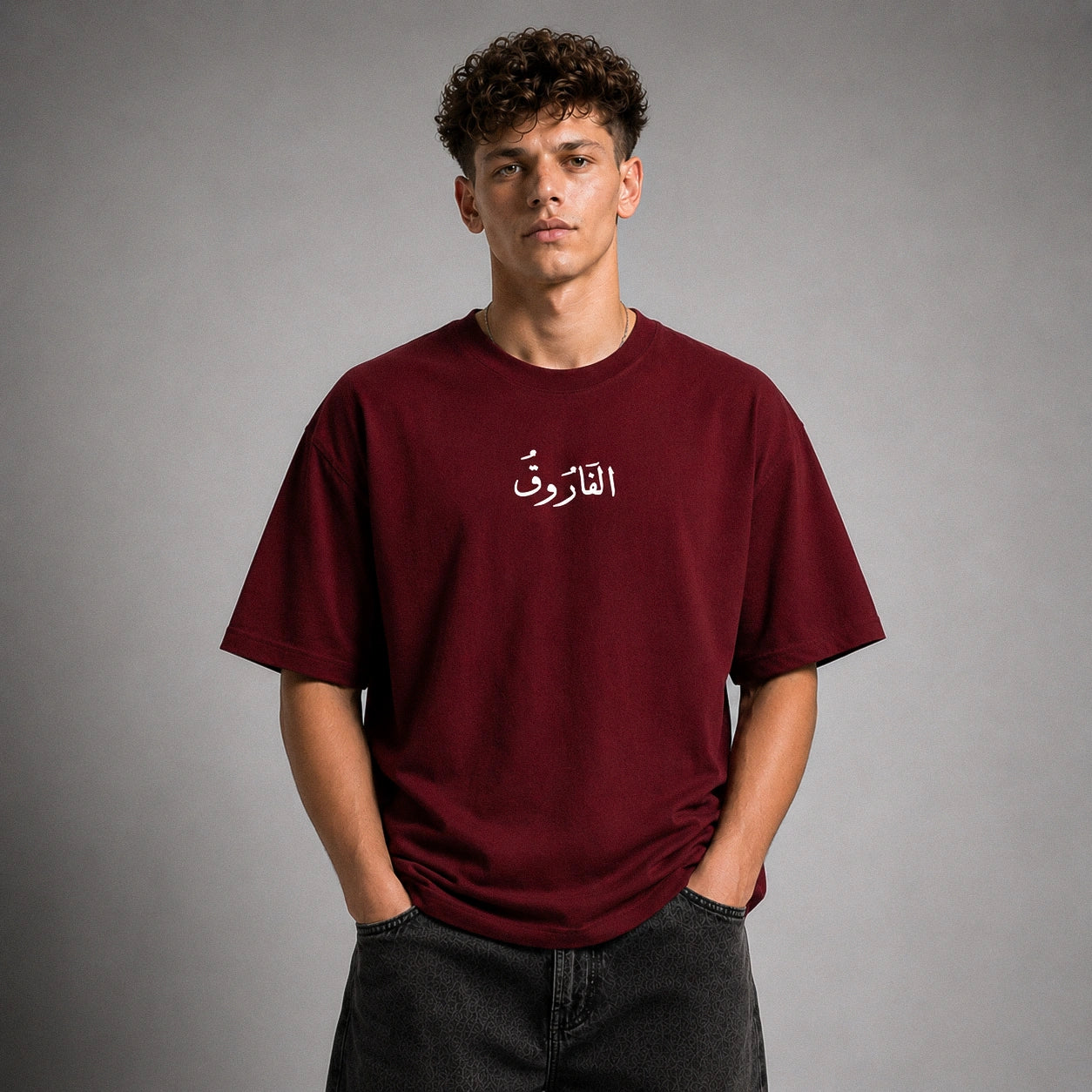 A young man with curly brown hair wearing an oversized maroon t-shirt with white Arabic calligraphy on the chest, paired with dark gray jeans, standing against a neutral gray studio background.