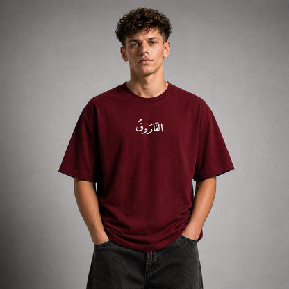 A young man with curly brown hair wearing an oversized maroon t-shirt with white Arabic calligraphy on the chest, paired with dark gray jeans, standing against a neutral gray studio background.
