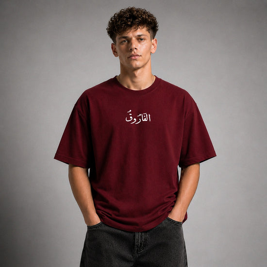 A young man with curly brown hair wearing an oversized maroon t-shirt with white Arabic calligraphy on the chest, paired with dark gray jeans, standing against a neutral gray studio background.