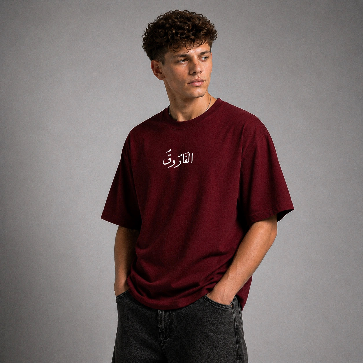 A young man with curly dark hair wearing an oversized maroon t-shirt with white Arabic calligraphy on the chest, paired with dark gray jeans, standing against a neutral gray studio background.