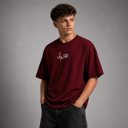 A young man with curly dark hair wearing an oversized maroon t-shirt with white Arabic calligraphy on the chest, paired with dark gray jeans, standing against a neutral gray studio background.