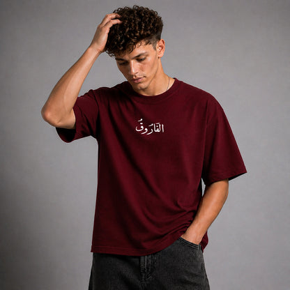 A man wearing a maroon crewneck t-shirt with white Arabic calligraphy on the chest, paired with dark gray jeans, posing against a solid gray studio background.