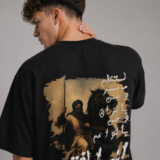 Person wearing a black t-shirt with a graphic design on the back, featuring a figure on horseback and Arabic text.