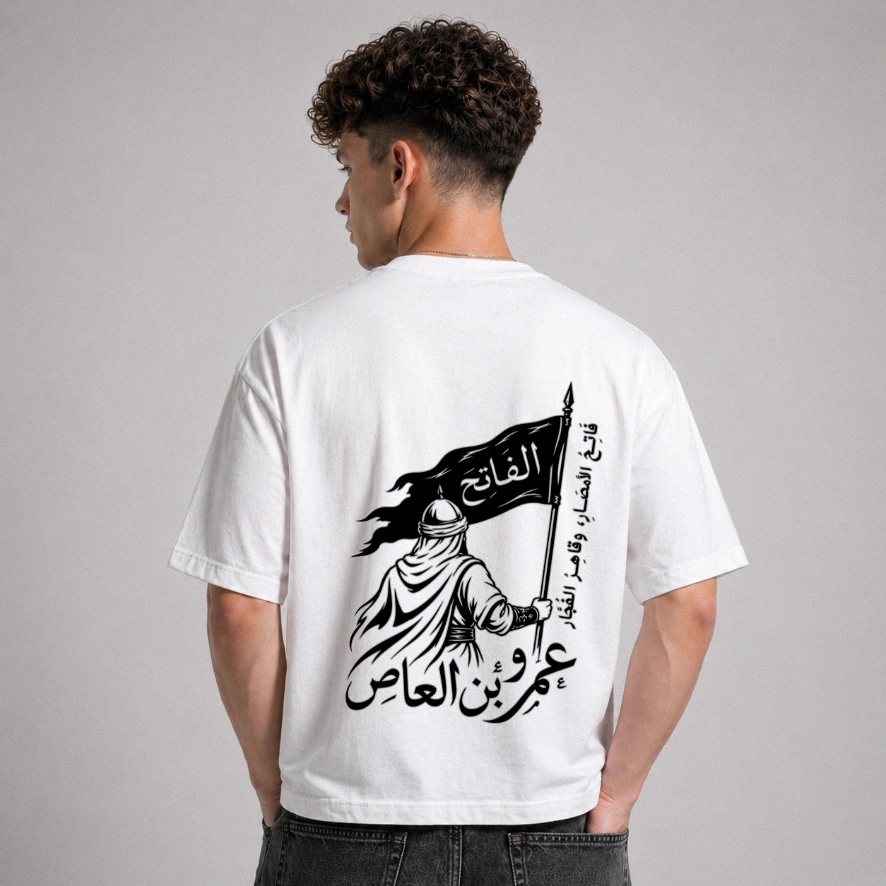 Person wearing a white t-shirt with a black graphic and Arabic text on the back, standing against a gray background.
