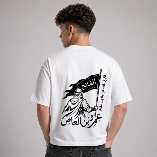 Person wearing a white t-shirt with a black graphic and Arabic text on the back, standing against a gray background.