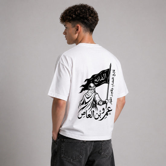 Person wearing a white t-shirt with Arabic text and graphic design on the back, standing against a gray background.