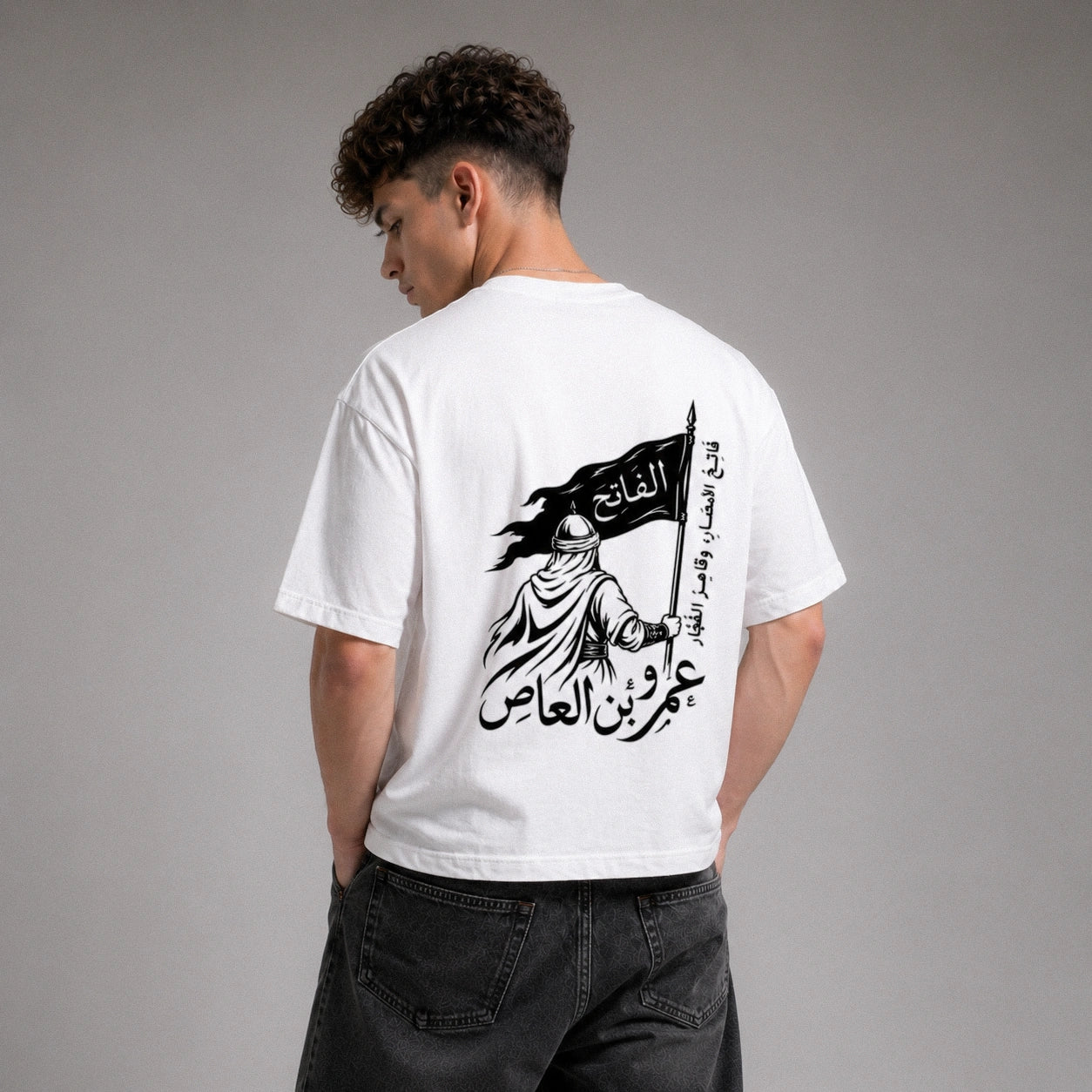 Person wearing a white t-shirt with Arabic text and design on the back, standing against a gray background.