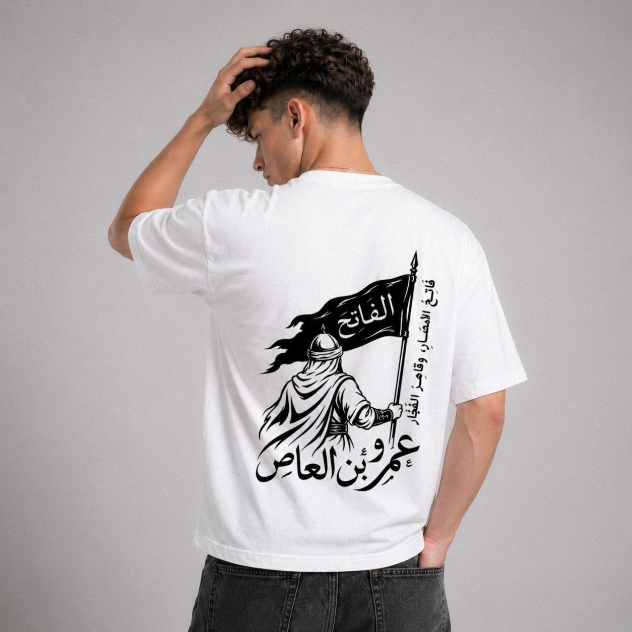 Person wearing a white t-shirt with Arabic text and graphic design on the back, standing against a gray background.