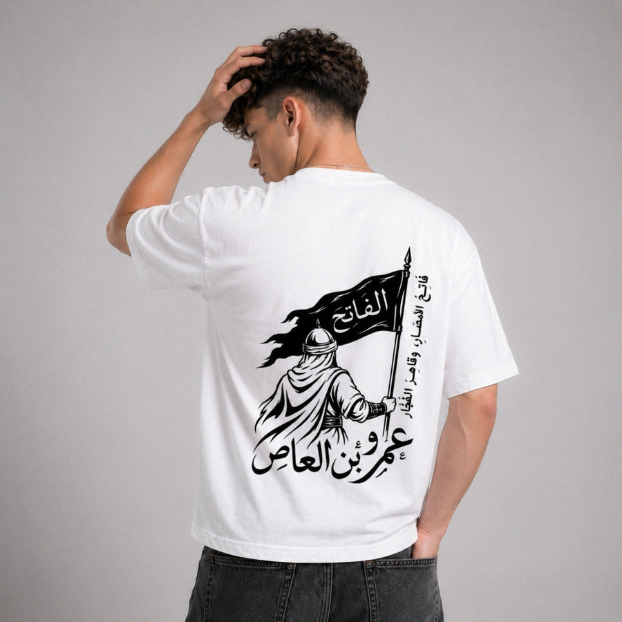 Person wearing a white t-shirt with Arabic text and graphic design on the back, standing against a gray background.