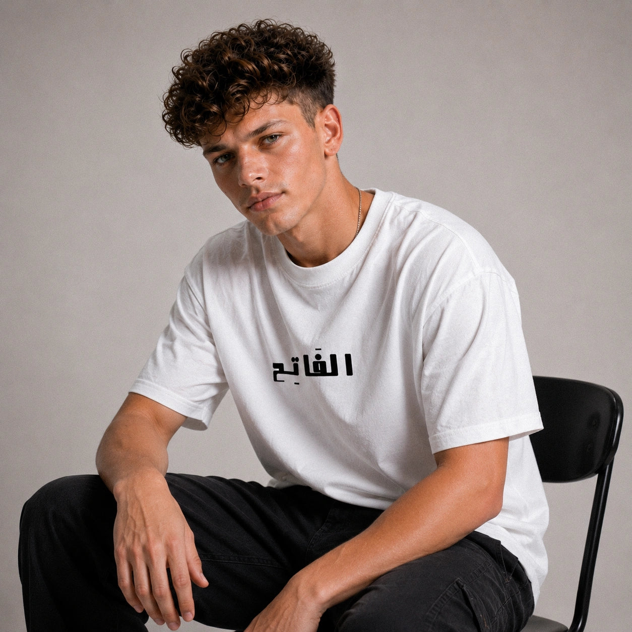 Person wearing a white t-shirt with Arabic text sitting on a chair against a plain background