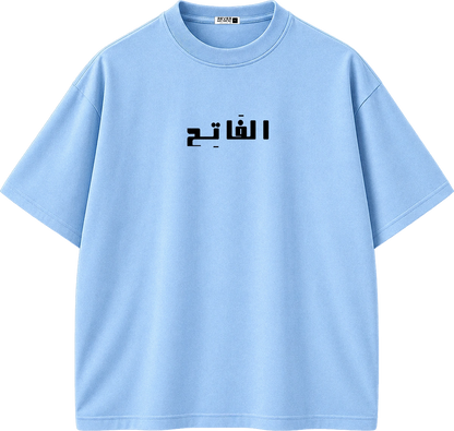 A light blue short-sleeve crewneck t-shirt with black Arabic text printed across the chest, displayed on a clean white background.