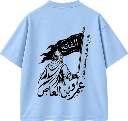 A light blue short-sleeve T-shirt shown from the back, featuring a large black graphic print of a robed figure holding a flag with Arabic text.
