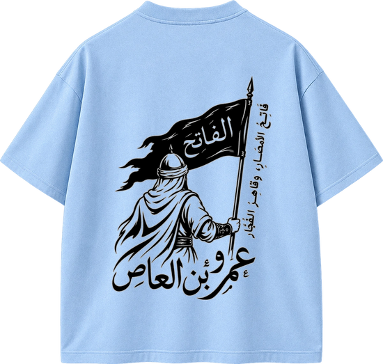 A light blue short-sleeve T-shirt shown from the back, featuring a large black graphic print of a robed figure holding a flag with Arabic text.