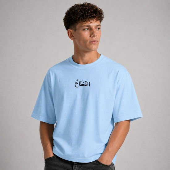 A man wearing a light blue crewneck t-shirt with black Arabic calligraphy on the chest, standing against a neutral gray studio background.