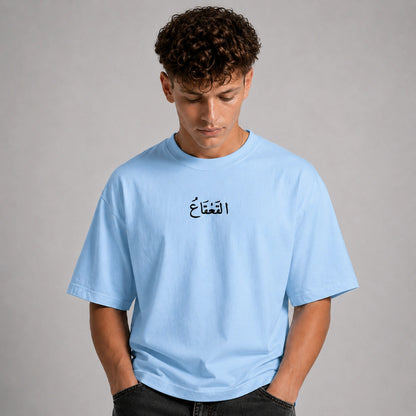 A man wearing a light blue crewneck t-shirt with black Arabic calligraphy printed on the chest, posed against a neutral gray studio background.
