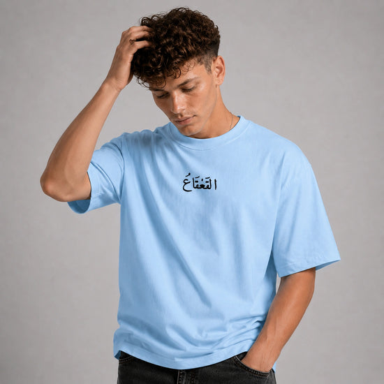 A man wearing a light blue crewneck t-shirt with black Arabic calligraphy on the chest, posed against a neutral gray studio background.