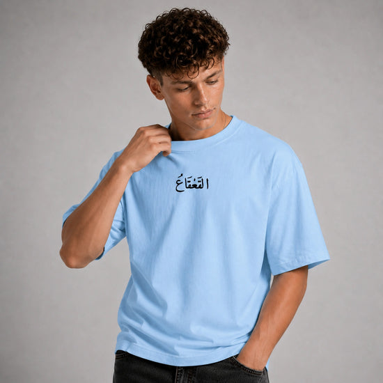 A young man with curly brown hair wearing a light blue crewneck t-shirt with black Arabic calligraphy on the chest, posed against a neutral gray studio background.