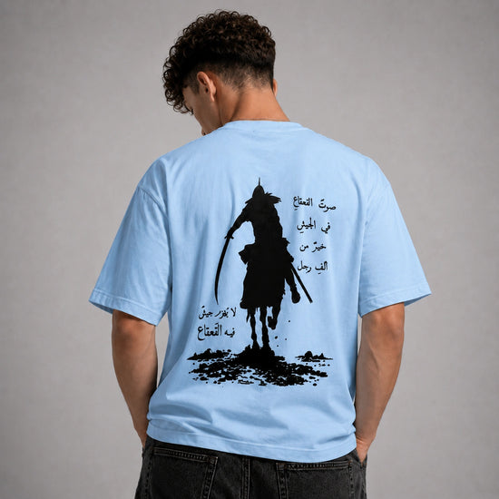 A man viewed from behind wearing a light blue t-shirt with a large black graphic print of a samurai silhouette and Arabic calligraphy on the back, set against a plain gray studio background.