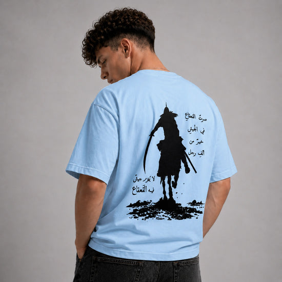 A man wearing a light blue t-shirt, viewed from the back. The shirt features a large black graphic of a samurai silhouette with Arabic calligraphy printed on the back.