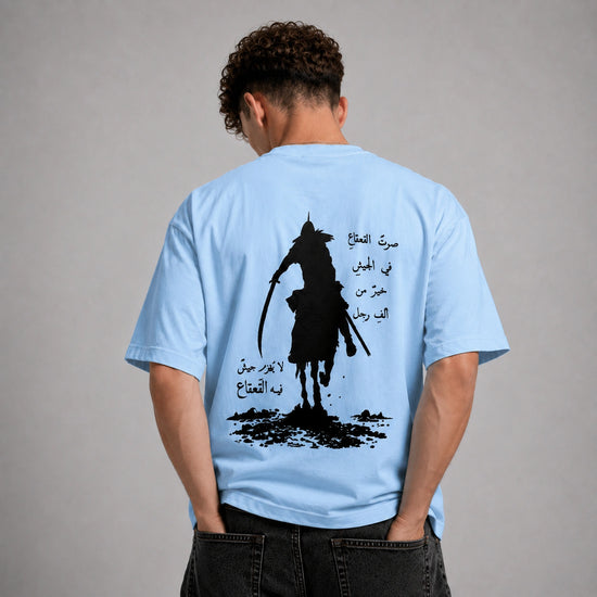 A man viewed from behind wearing a light blue t-shirt with a large black graphic print on the back, paired with dark gray jeans, against a neutral gray background.