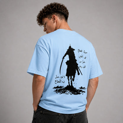 A man wearing a light blue t-shirt, viewed from behind, showing a large black graphic print of a samurai on horseback with Arabic text on the back.