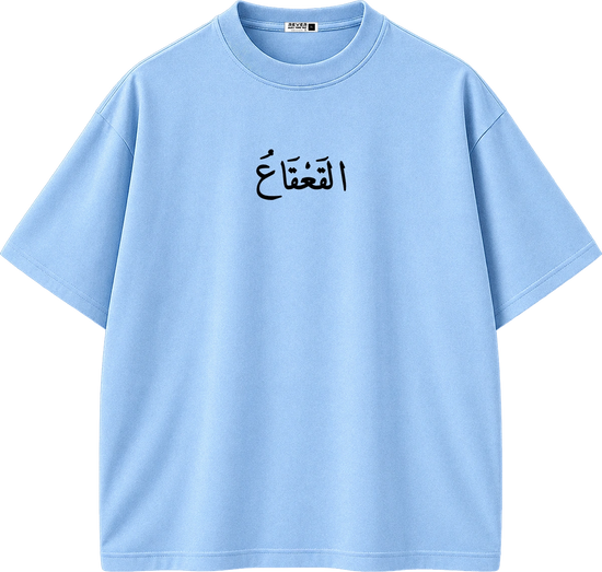 A light blue short-sleeve crewneck t-shirt with black Arabic calligraphy printed on the center chest, displayed on a clean white background.