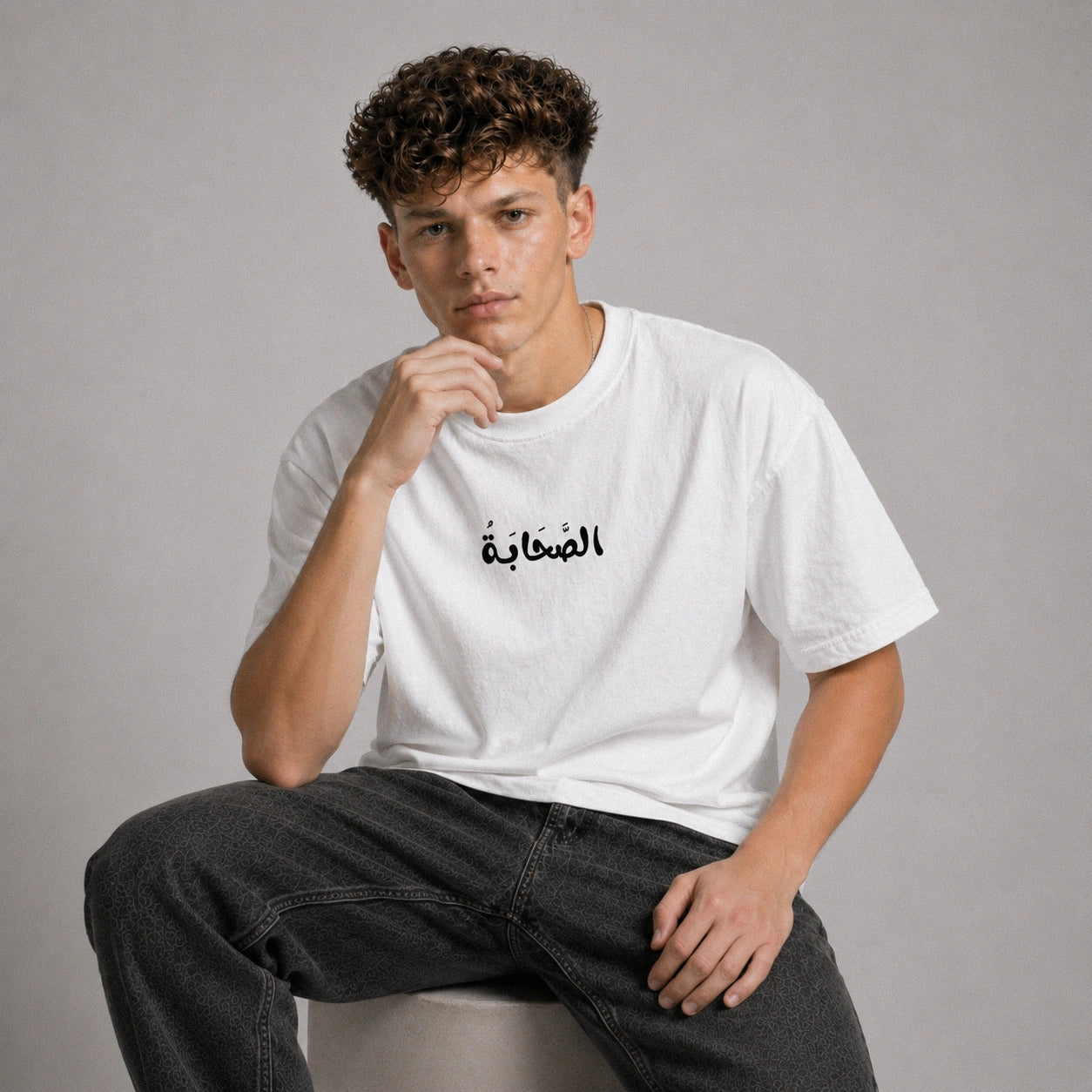 Person wearing a white t-shirt with Arabic text sitting on a gray background