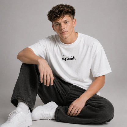 Person wearing a white t-shirt with Arabic text, sitting on a gray background