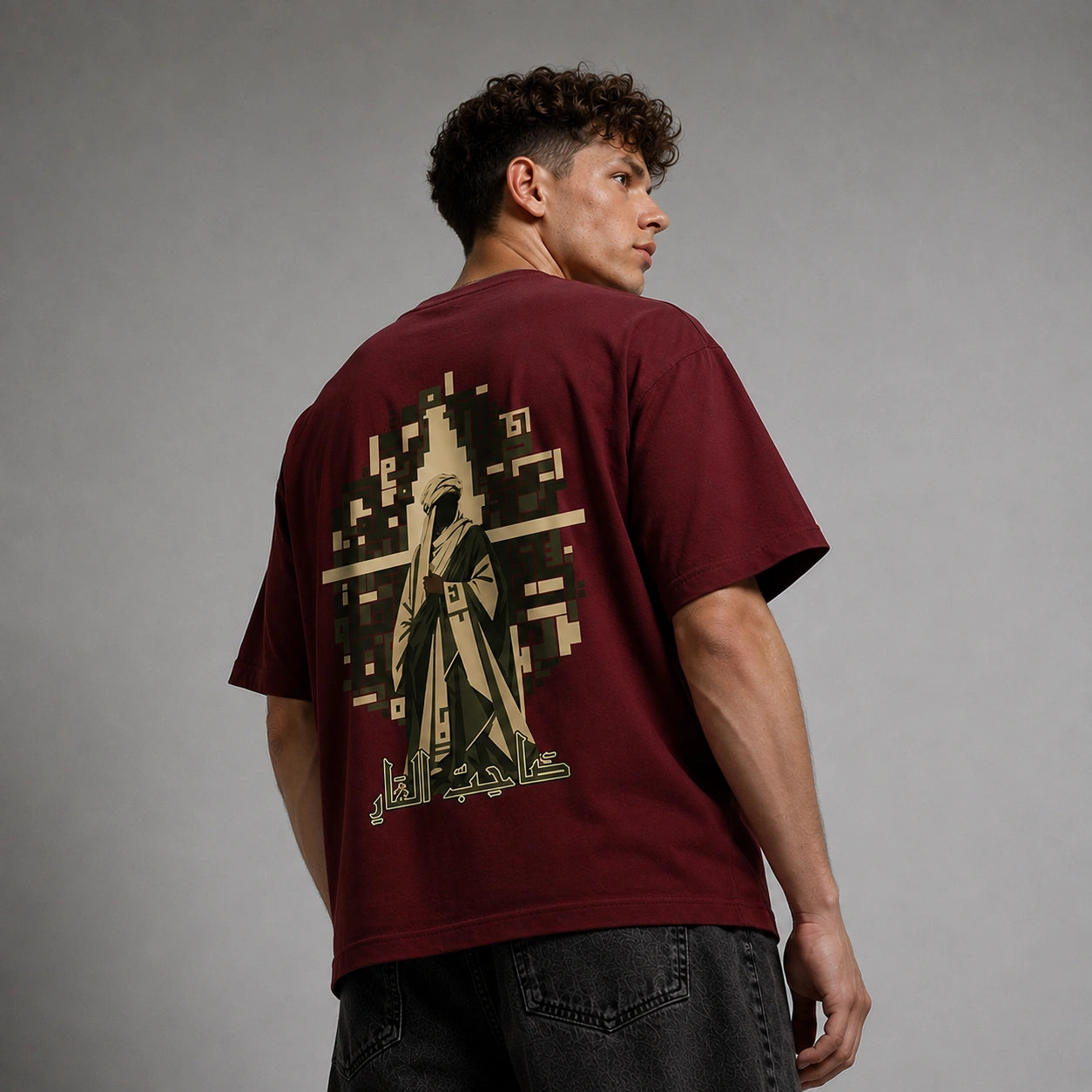 A man wearing a maroon t-shirt with a large beige graphic print on the back, shown from behind against a gray studio background.