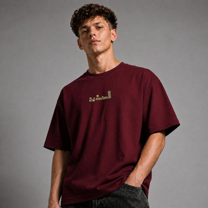 A young man with curly brown hair wearing an oversized maroon t-shirt with gold Arabic calligraphy on the chest, paired with dark gray jeans, photographed against a neutral gray studio background.