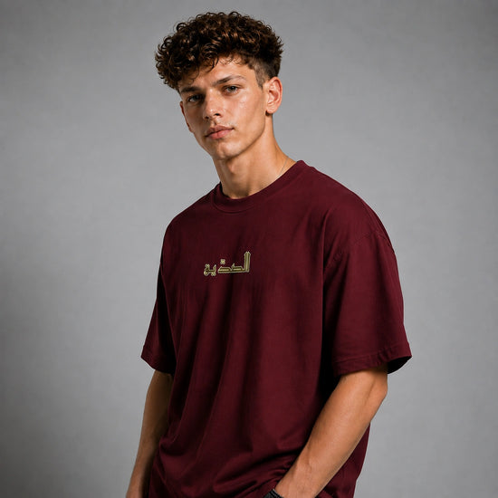 A young man with curly brown hair wearing a maroon crewneck t-shirt with gold Arabic calligraphy on the chest, photographed against a neutral gray studio background.