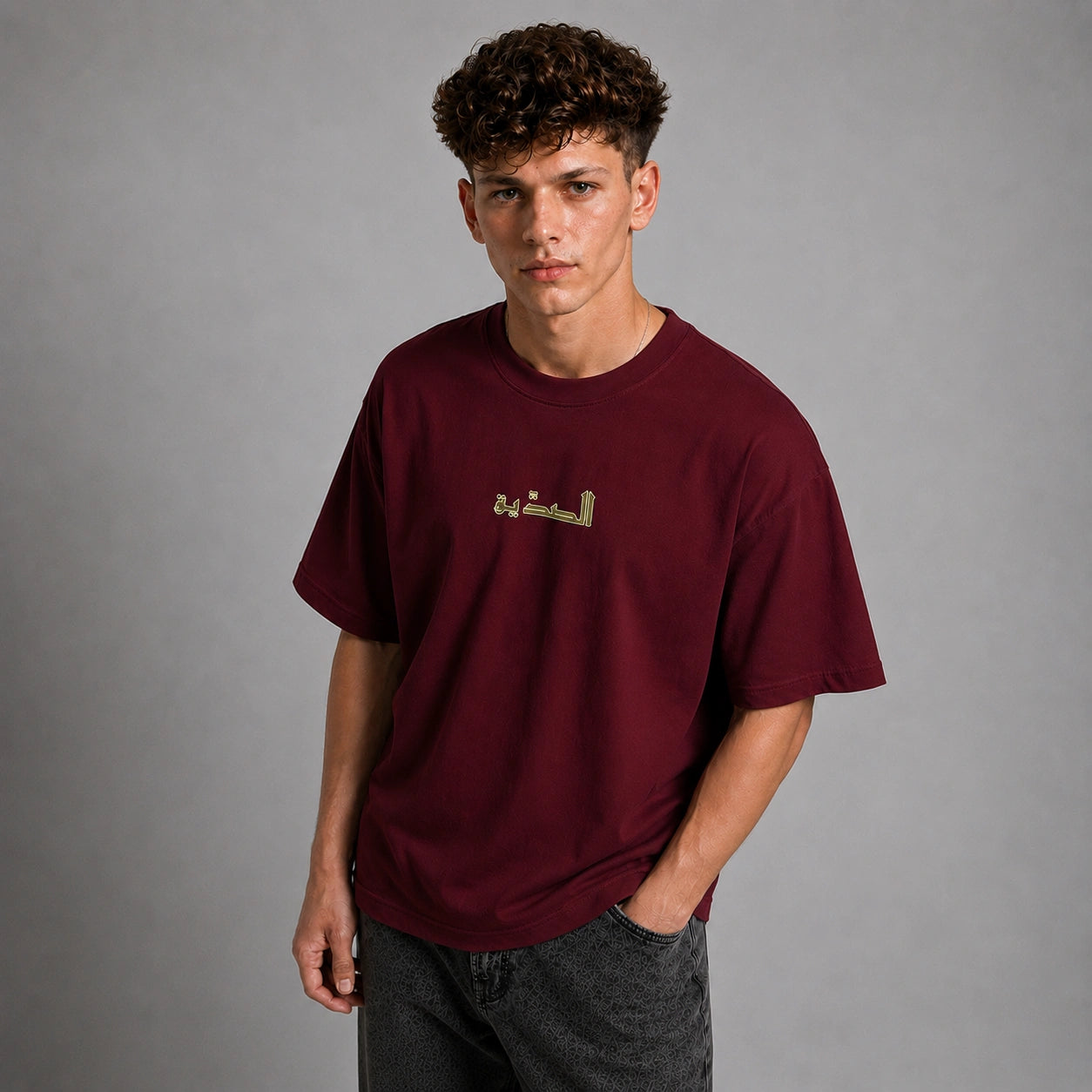 A young man with curly brown hair wearing a maroon crewneck t-shirt with gold Arabic calligraphy on the chest, paired with dark gray jeans, standing against a neutral gray studio background.