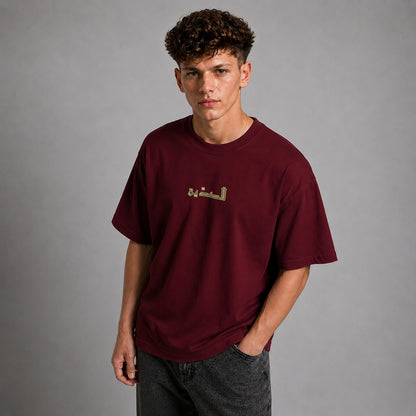 A young man with curly brown hair wearing a maroon crewneck t-shirt with gold Arabic calligraphy on the chest, paired with dark gray jeans, standing against a neutral gray studio background.