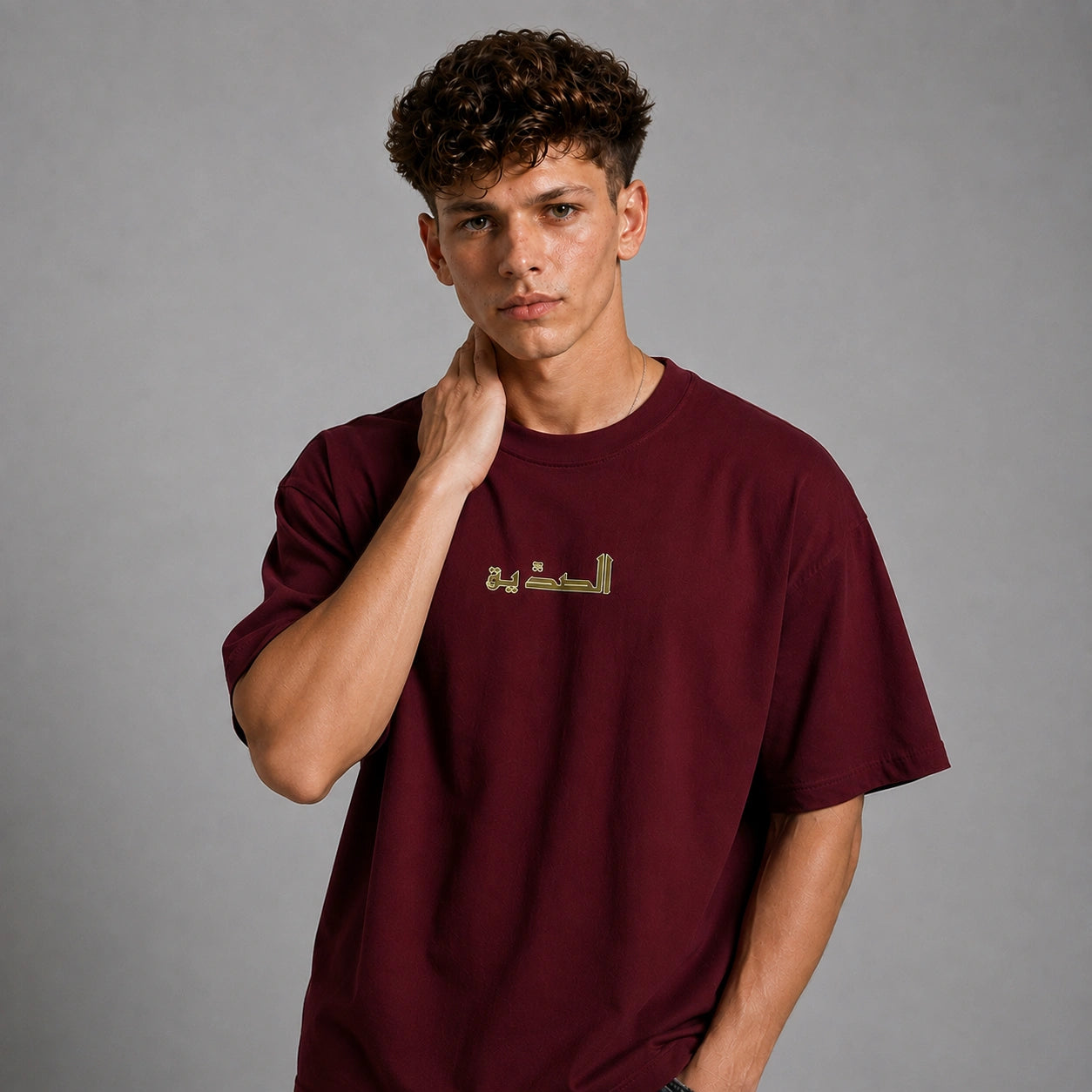 A young man with curly brown hair wearing a maroon crewneck t-shirt with gold Arabic calligraphy on the chest, posing against a solid gray studio background.