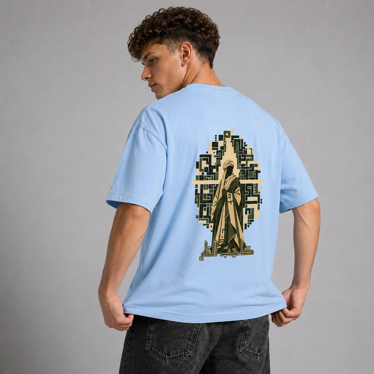 A man wearing a light blue t-shirt with a large graphic print on the back, shown from behind against a neutral gray studio background.
