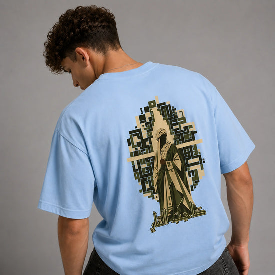 A man wearing a light blue t-shirt, viewed from behind, showcasing a large graphic print on the back. The graphic features a stylized figure in a robe integrated with a pixelated geometric cross and Arabic calligraphy at the bottom, set against a solid gray studio background.
