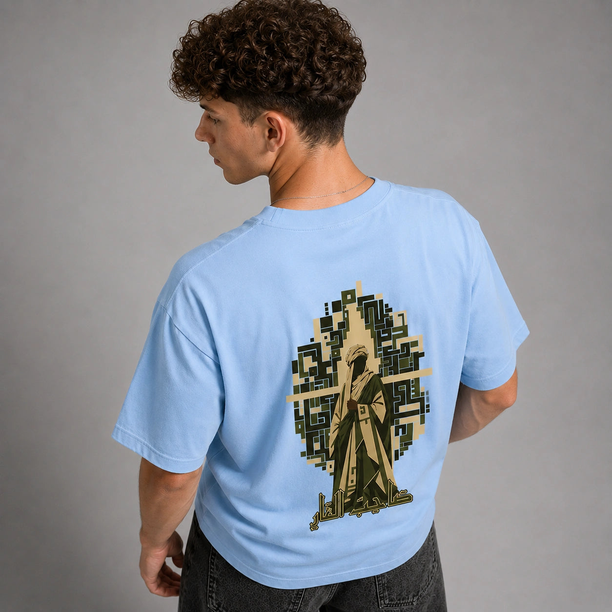 A man wearing a light blue t-shirt, viewed from the back. The shirt features a large graphic print of a hooded figure in a trench coat set against a pixelated tree silhouette.