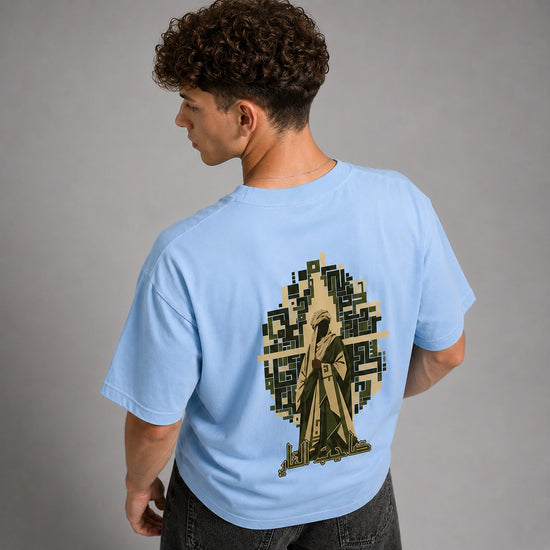 A man wearing a light blue t-shirt, viewed from the back. The shirt features a large graphic print of a hooded figure in a trench coat set against a pixelated tree silhouette.
