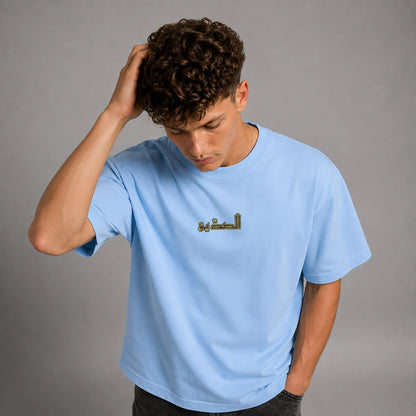 A man with curly brown hair wearing a light blue crewneck t-shirt with gold Arabic calligraphy on the chest, posing against a neutral gray background.